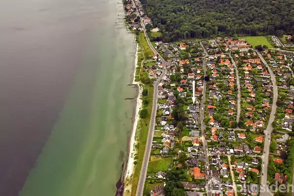 Kystlinje løber parallelt med en vej, der adskiller en stribe skov og beboelse med mange huse. Vandet er grønt nær kysten og mørkere længere ud.
