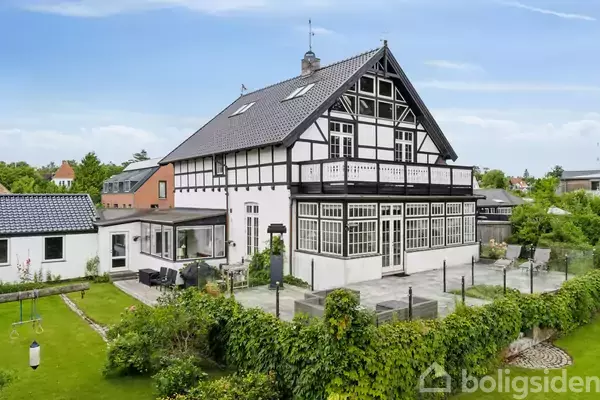 Et stort, klassisk bindingsværkshus med sort-hvid facade står på en velplejet havegrund. Solsengen står på den stenbelagte terrasse omgivet af grønne buske og blomsterbede under en blå himmel.