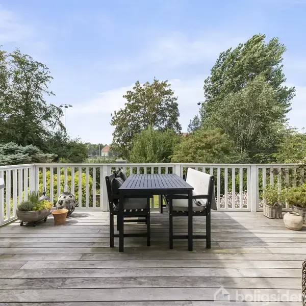 Bord og stole står på en træterrasse, omkranset af hvide gelændere. Terrassen er dekoreret med potteplanter, og i baggrunden ses en have med træer og buske under en blå himmel.