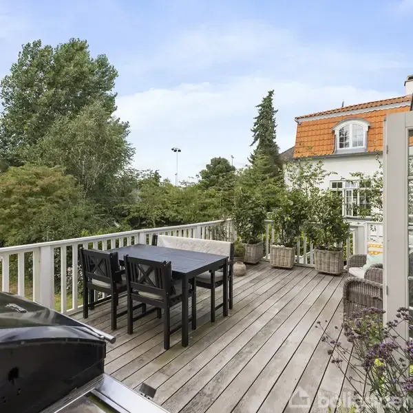Træterrasse med et bord og fire stole står udendørs. Grillen er placeret i forgrunden. Omgivet af grønne træer og en hvid bygning med orange tag.