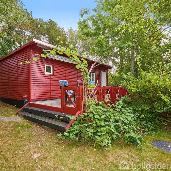 Rødt træhus med terrasse, omkranset af frodig vegetation og høje træer. Trappen fører op til en lille veranda med stole, mens grønne buske vokser tæt på huset.