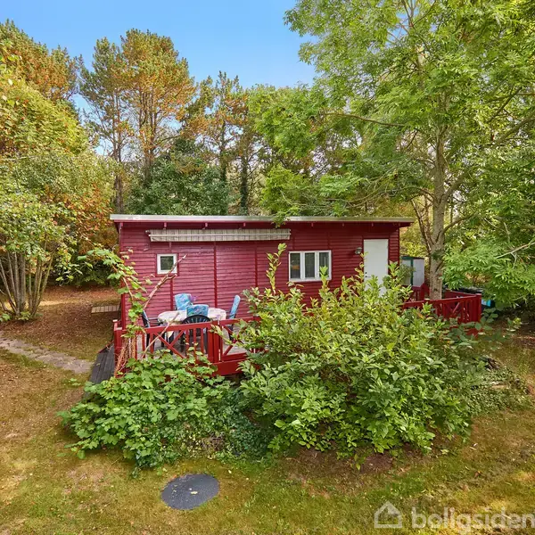 Rødt træhus står stille omgivet af frodig vegetation og store træer; en lille terrasse foran huset rummer blå stole, der skaber en hyggelig atmosfære i den grønne have.