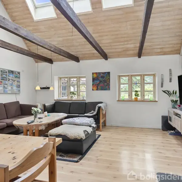 Stue med brun sofa arrangeret omkring et træbord. TV står på et hvidt møbel med dekorationer. En pejs i hjørnet. Blomster i vase på et spisebord. Vinduer og ovenlys med trærammer.