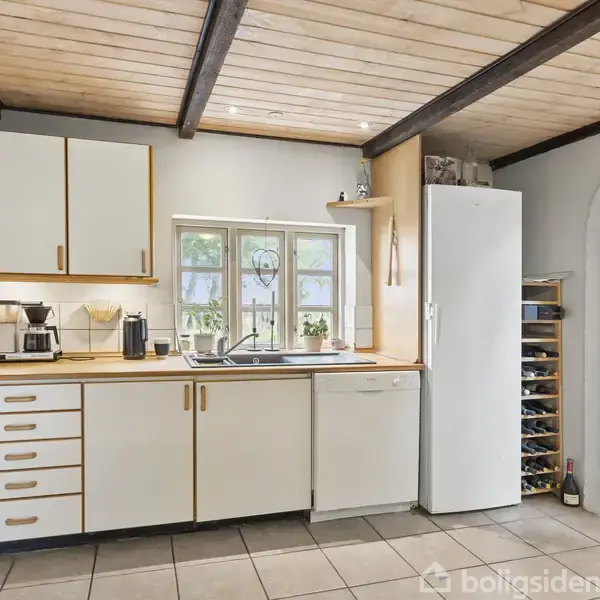 Et køkken med hvide skabe og skuffer, en vask under et vindue, og en høj, hvid køleskab ved siden af. Kakkelgulv, træloft med synlige bjælker.