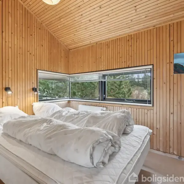 En dobbeltseng med hvide dyner står i et træbeklædt soveværelse. Store vinduer giver udsigt til grønne træer udenfor. En lille maleri hænger på væggen.