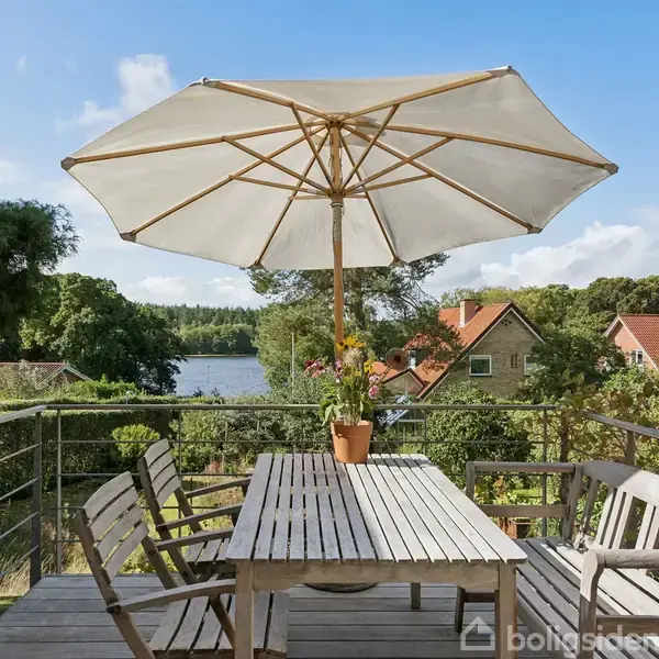 Parasol skygger over et træbord med stole på en terrasse. En blomsterpotte står på bordet. Baggrunden omfatter en have, stuele af huse og en sø i det fjerne.