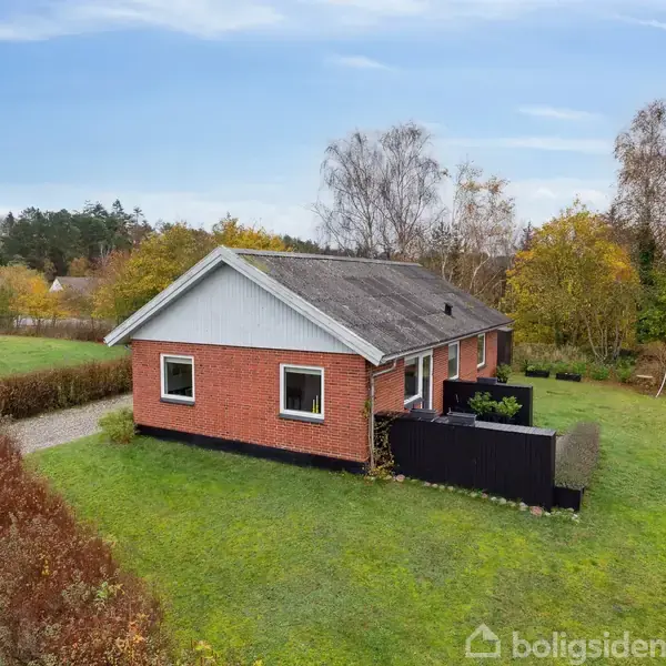 Et rødt murstenshus med sort skur omkranset af grønne træer og buske står ved en grusvej. Huset har en græsplæne foran og er omgivet af hække.