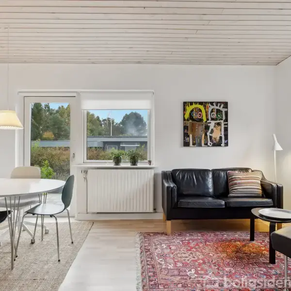 Sort lædersofa står i moderne stue med lyse vægge og trægulv. Rundt spisebord med stole er til venstre; vindue med udsigt over have til højre. Ingen tekst fundet.