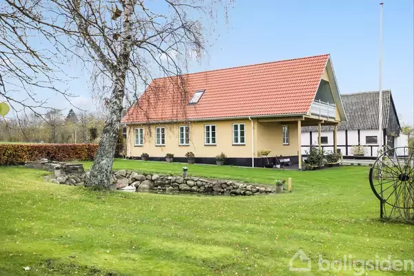 Et gult hus med rødt tag står på en velplejet græsplæne. Ved siden af er der en stengærde og et bladløst træ. Bagved ses et andet hus.