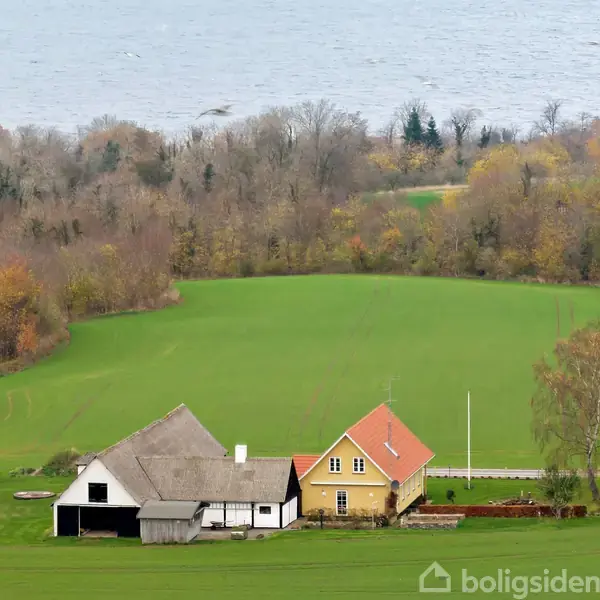 To huse ligger ved en grøn mark, omgivet af træer i efterårsnuancer. I baggrunden ses en sø eller hav, der giver en naturskøn udsigt.