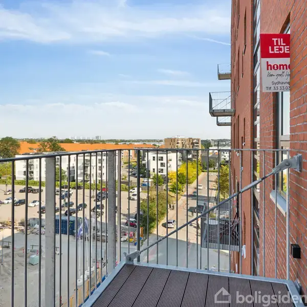Balkon med metalrækværk hænger ud fra en murstensbygning. I baggrunden ses en byudsigt med parkeringspladser og bygninger. Skilt med teksten "TIL LEJE" hænger på væggen.