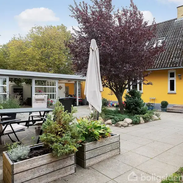 En gårdhave med havemøbler og plantekasser. Et træ står i midten. Gårdhaven omgives af et gult hus med hvide vinduer og dør. Der er også en carport med glaspartier.