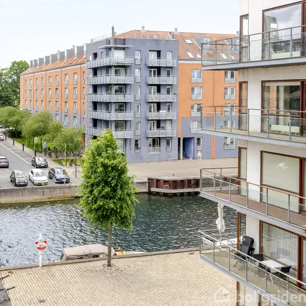 Bygninger med store vinduer står ved en kanal. Flere etager har balkoner. Gade med parkerede biler ses i baggrunden nær et rødt og et blåt lejlighedskompleks. En trædækket terrasse ses nærmest.