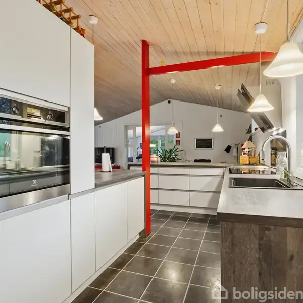 Et moderne køkken med hvide skabe og indbygget ovn. Står i et rum med træloft, rødt stolpeelement, store vinduer og lamper, der hænger fra loftet.