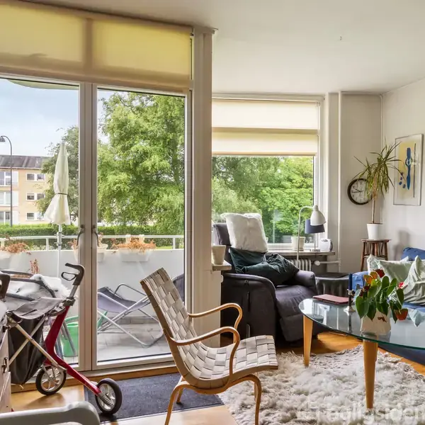 Stue med blå sofa, glasbord og stole i et veloplyst rum. Store vinduer viser en balkon med planter udenfor og udsigt til en grøn park med træer. Malerier på væggen.