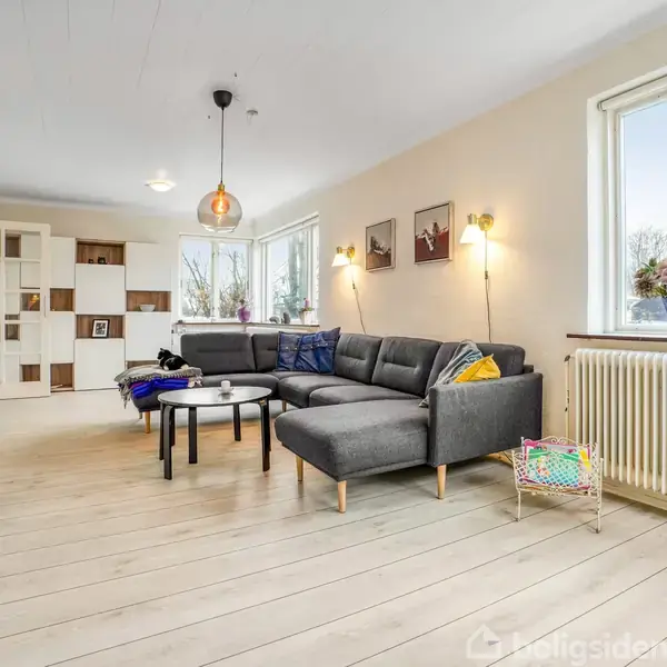 Sofa placeret mod væg, flankeret af små borde. Rum med trægulv, vægdekorationer og store vinduer med udsigt udendørs. Lamper skaber varm belysning.