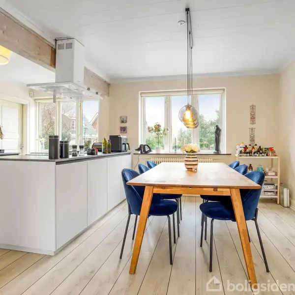 Et spisebord med blå stole står i et lyst køkken med trægulve. Køkkenøen har hvide skabe, og store vinduer giver naturligt lys. Billeder hænger på væggen.