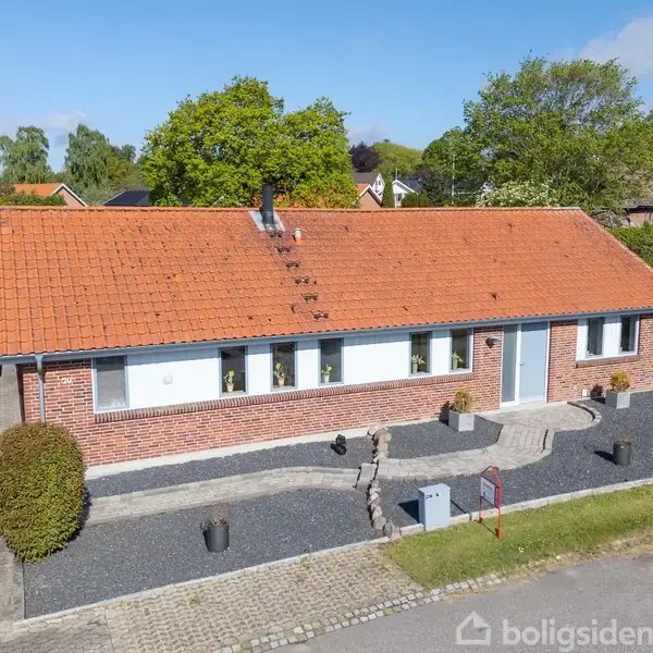 Rødt murstenshus med rødt tegltag, beliggende i en velplejet have med grusbelagt forhave, planter og flisebelægning. En carport med en bil er synlig til venstre.