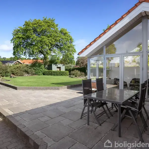 Et havebord med fire stole står på en flisebelagt terrasse. I baggrunden ses en grøn have med træer og buske under en klar blå himmel.
