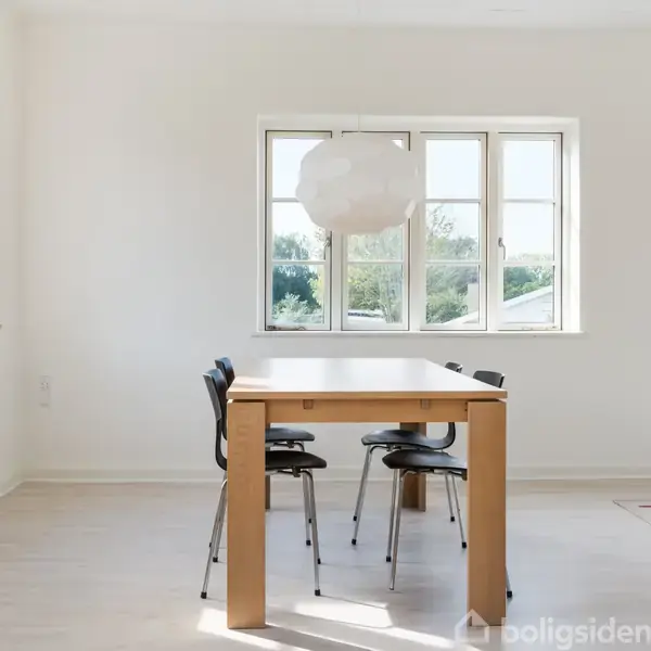 Et spisebord med fire sorte stole står i et lyst rum. En stor lampe hænger over bordet. Vinduer i baggrunden lader sollys trænge ind, belyst af udsigt til træer.