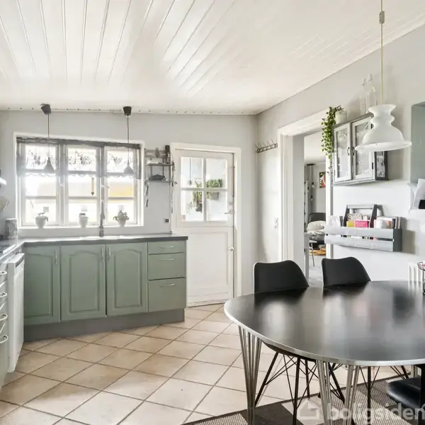 Køkkenbord med sorte stole står i midten. Der er grå skabe og hvide fliser, en vask under vinduet, og åbne hylder med dekorationer. Lysekrone hænger fra loftet.