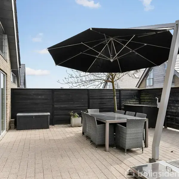 En terrasse med et stort, sort parasol over et spisebord og seks stole. Omgivet af træhegn, med nogle planter i krukker og en bygning til venstre.
