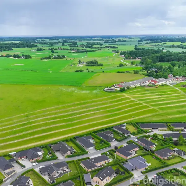 Huse ligger i en planlagt bebyggelse omgivet af grønne marker. I baggrunden ses skovområder og en lille landsby. Himlen er overskyet med få lyse pletter.