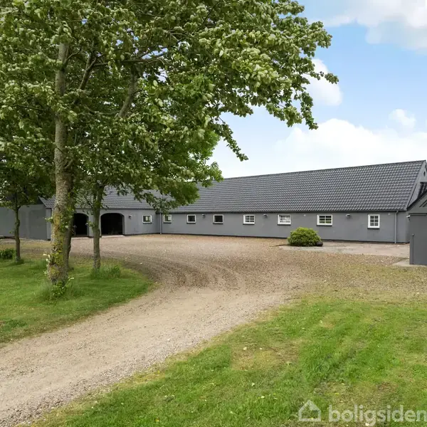 En grå, aflång bygning står stille i et åbent gårdmiljø, omgivet af en grusvej og grønne træer. Himmel med skyer kan ses i baggrunden.