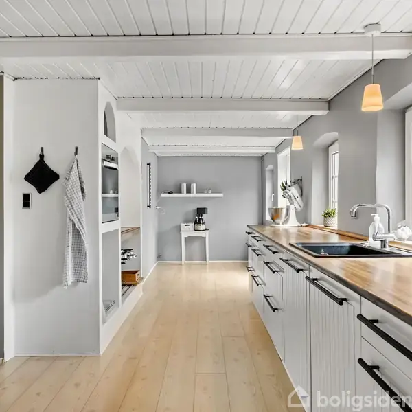 Et moderne køkken med hvide skabe og træbordplade, hvor en vask er integreret. Store vinduer giver masser af lys. En hylde med køkkenartikler ses længere inde i rummet.