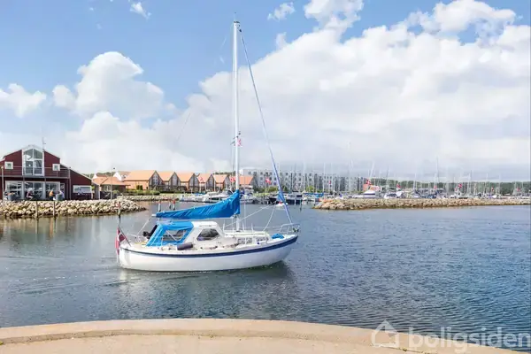 En sejlbåd med blå presenning flyder stille i en havn med pælehuse i baggrunden og en let overskyet himmel ovenover.