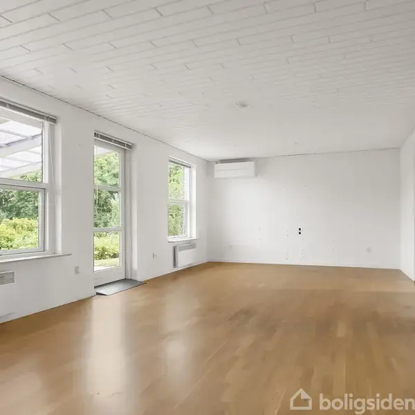 Et rummeligt stueområde med trægulv, store vinduer og hvide vægge. Rummet har indbygget aircondition, og vinduerne viser ud til en frodig grøn have.