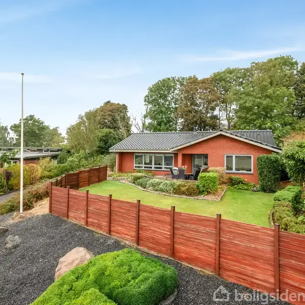 Rødt murstenshus står stille omkranset af en velplejet have med en træhegn. Det ligger i et grønt boligområde, omgivet af træer og en blå himmel i baggrunden.