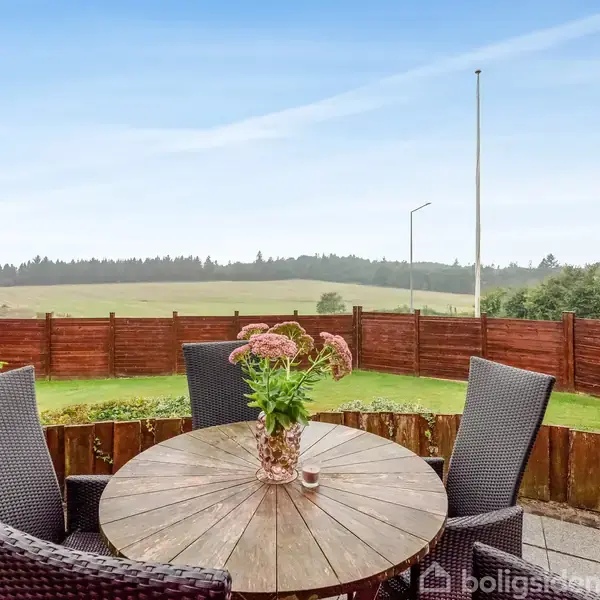 Rundt havebord med flettede stole står på terrasse, dekoreret med blomster i vase. Baggrund viser græsplæne og åbent landskab med træer og blå himmel.