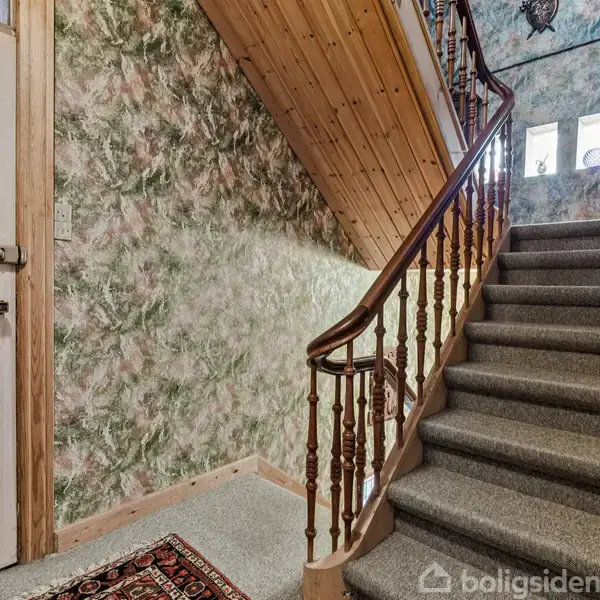 Trappeopgang med tæppebelagte trin og trægelænder fører op til første sal. Væggene har et blomstret mønster, og der er en trædør til venstre i en indgangshal.