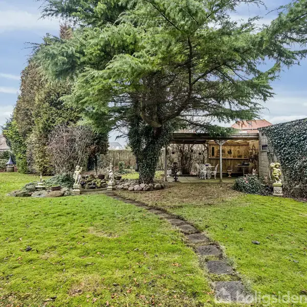 En stor, grøn have med græsplæne og en flisebelagt sti fører til et overdækket område med borde og stole omgivet af høje træer og en hæk i baggrunden.