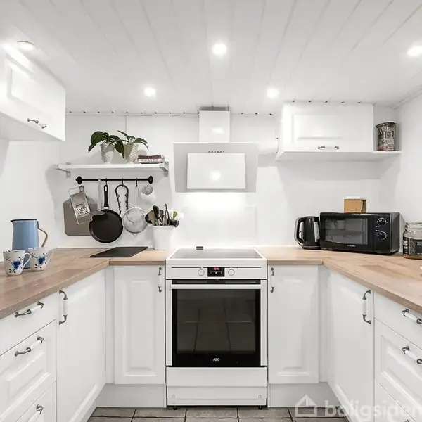 Et moderne køkken viser ovn omgivet af hvide skabe og træbordplader. En emhætte hænger over ovnen. Til venstre står en elkedel, til højre en mikrobølgeovn og frugt.