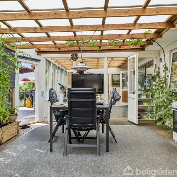 Et udendørs spisebord med stole står under en træpergola. Bredåbne glasdøre fører til en indendørs opholdsstue. Klatreplanter og potter tilføjer grønne nuancer rundt om området.