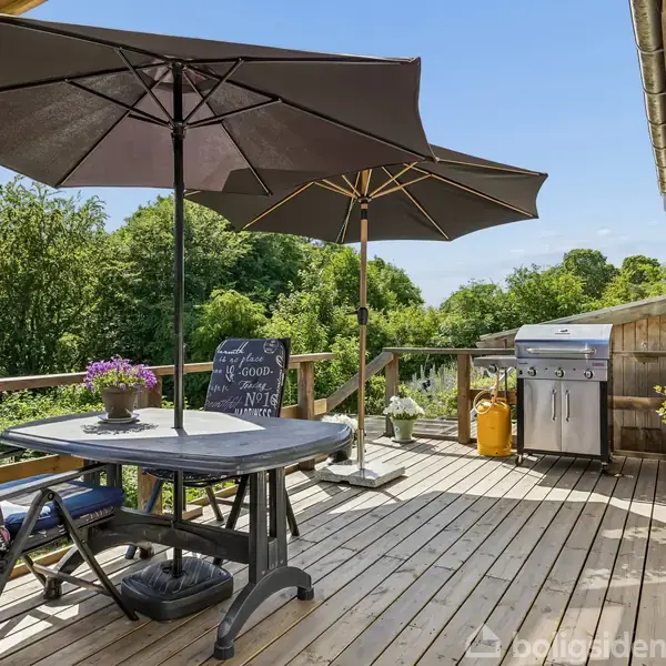 Terrasse med trædæk har to parasoller, bord og stole. På bordet står en krukke med blomster. Gasgrill og planter i potter langs rækværket. Omgivet af grøn natur.