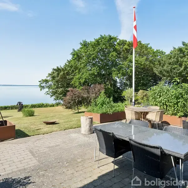 Et stort bord med stole står på en terrasse, omgivet af frodig have. I baggrunden ses en flagstang med Dannebrog og udsigt over fjorden.