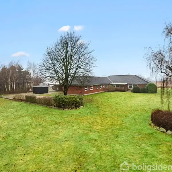Huset er en enkeltstående bygning med røde mursten og sort tag omgivet af en stor grøn have med træer og buske. Det er placeret i et åbent grønne område.