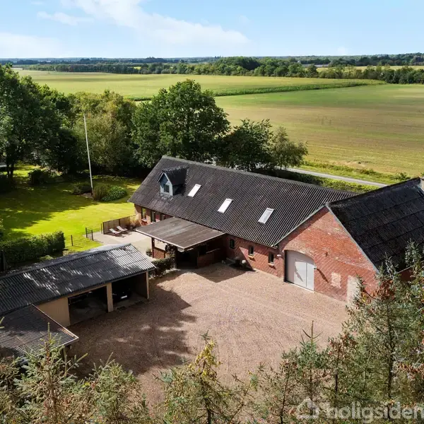 En stor, rød murstensbygning med sort tag er placeret i en grøn, landlig omgivelser. Ved siden af bygningen er der en åben garage. Landskabet er fladt med marker og træer.