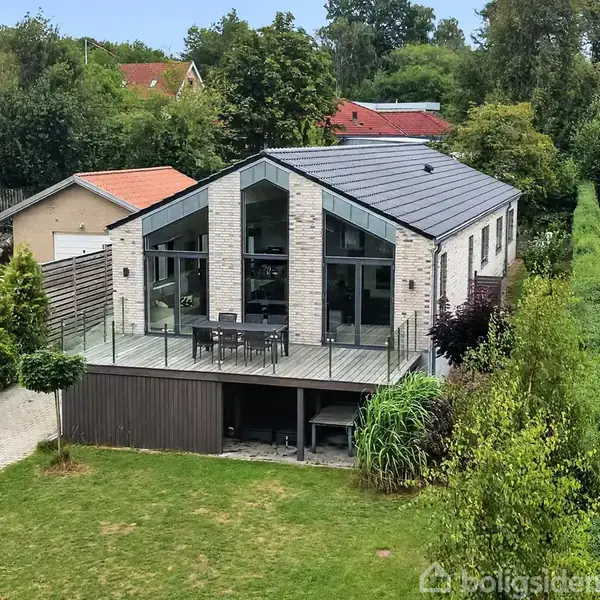 Et moderne hus med store glaspartier og valmtag har en rummelig terrasse med havemøbler. Det er omgivet af en velholdt have med en flisebelagt sti og tæt beplantning.