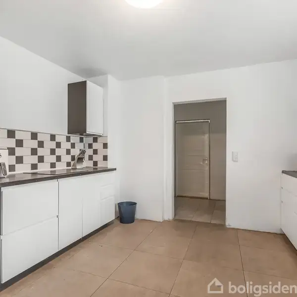 Køkken med hvide skabe og skuffer, har en indbygget ovn og mikrobølgeovn til venstre. Flisebelagt backsplash i sort og hvid. Rum er rummeligt med beige gulvfliser. Ingen tekst fundet.