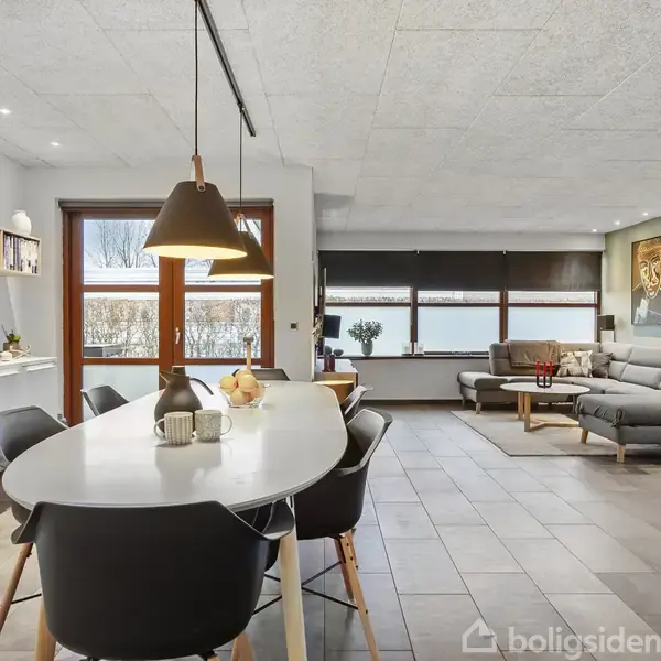 Spisebord med sorte stole står i et åbent, moderne stue-område. Bordet er dækket med en vase og en skål. Baggrunden viser en stor sofa og kunst på væggene.