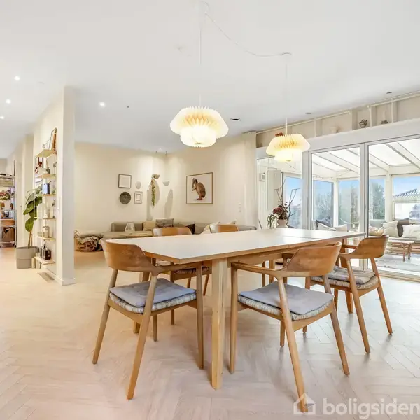 Et lyst spisebord med seks stole står i et åbent, veloplyst rum med trægulve. Store vinduer i baggrunden viser en udestue med udsigt til en have.