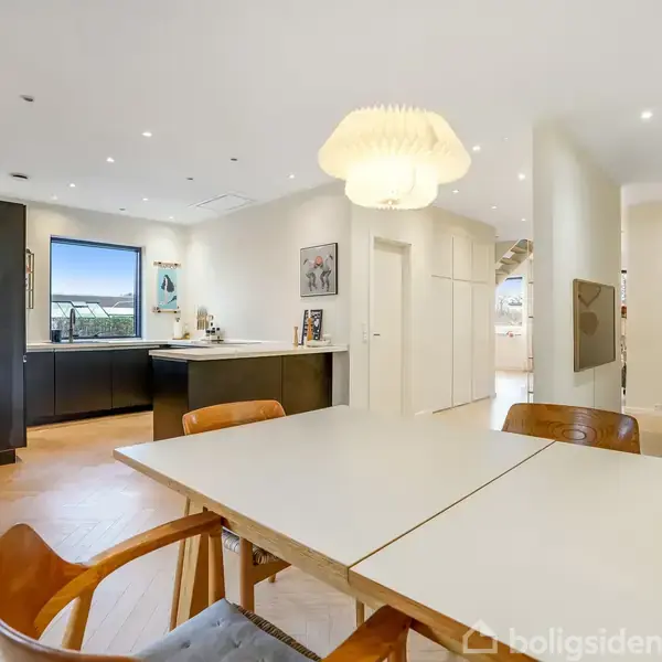 Et spisebord med træstole står i et lyst køkken-alrum. En moderne lampe hænger over bordet. Køkkenet har sorte skabe, hvid bordplade og et stort vindue.