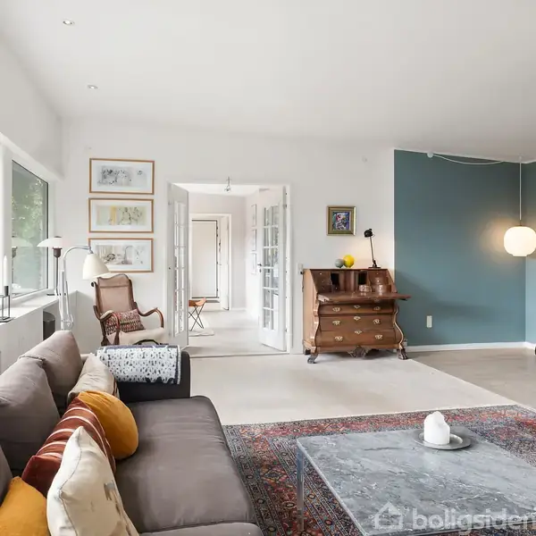 Sofa beliggende i stue foran stort vindue med udsigt til træer. Stue med pejs, indrettet med tæppe, bord, kommode og polstrede stole. Rummeligt med lyse vægge og kunst på væggene.