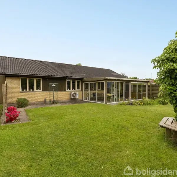 En villa med mørkt tag og gul murstensfacade, der står i en grøn have med velholdt græsplæne. Der er en glasudestue og blomster ved siden af huset.