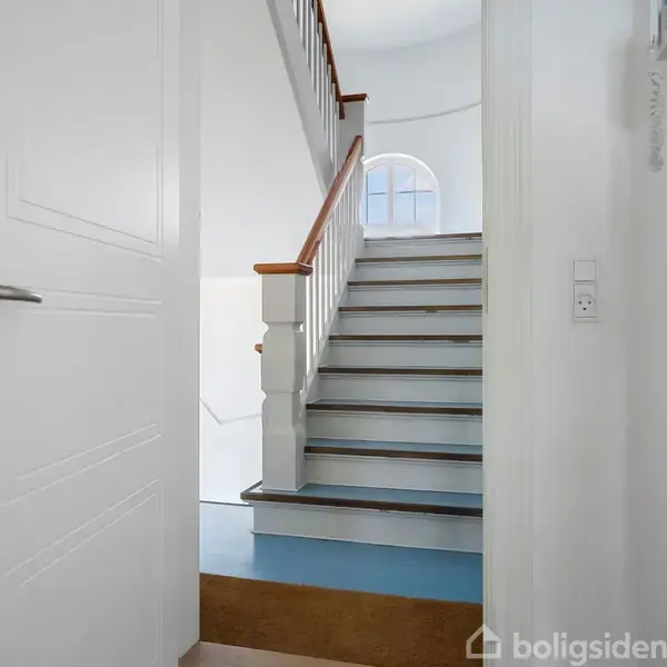 En hvid dør står åben og fører ind til en trappeopgang. Trappen har hvide trin med en trægelænder. Et rundt vindue er synligt for enden af trappen.