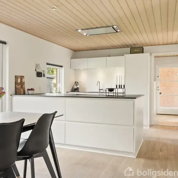 Et moderne, lyst køkken med en central køkkenø. To sorte stole står ved et spisebord med blomster. Træloft og lyse vægge skaber en varm atmosfære.